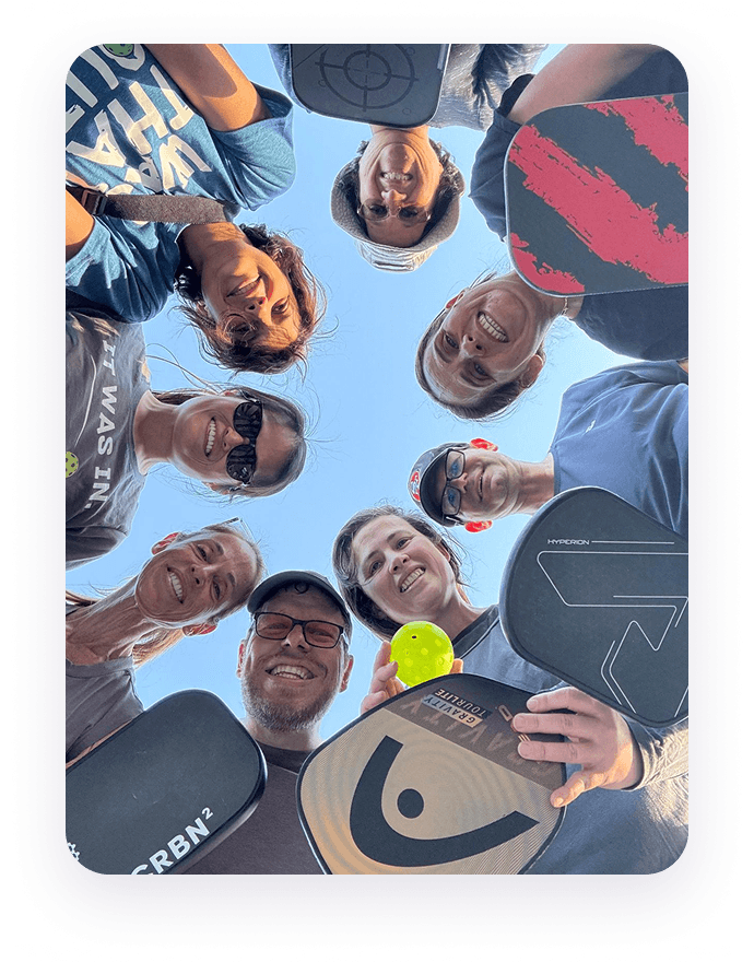 A group gathered around the camera outdoors, smiling with their pickleball paddles
